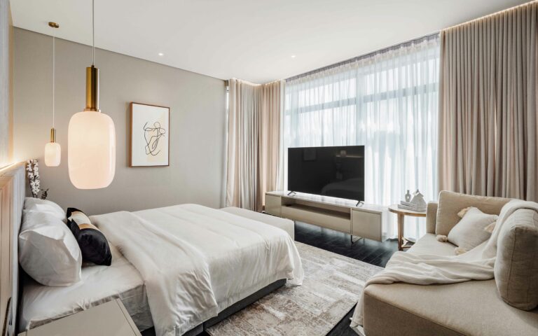 Experience the epitome of luxury in this spacious, well-lit bedroom featuring a plush queen-size bed, bedside sofa, and a flat-screen TV, all set against a backdrop of stunning interior design elements including expensive carpeting.