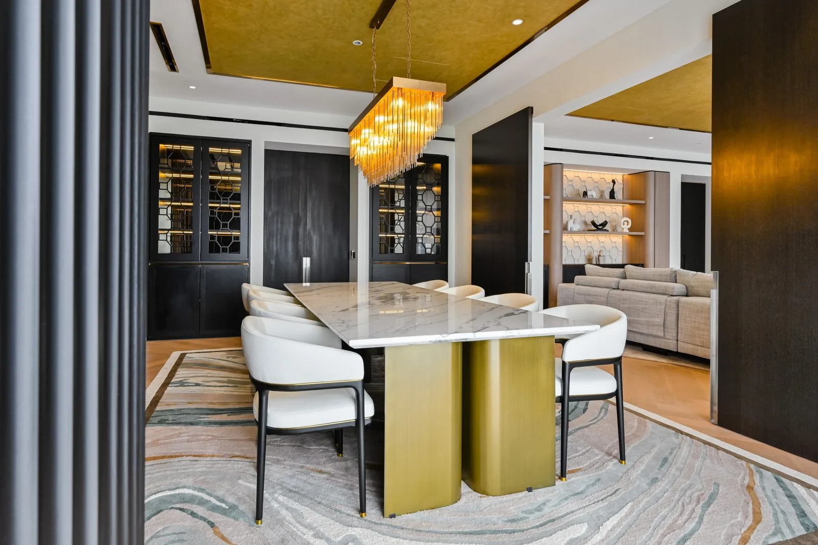 Modern dining room with a marble table on a gold base, white chairs and a linear crystal chandelier under a gold leaf ceiling at Aira Residence