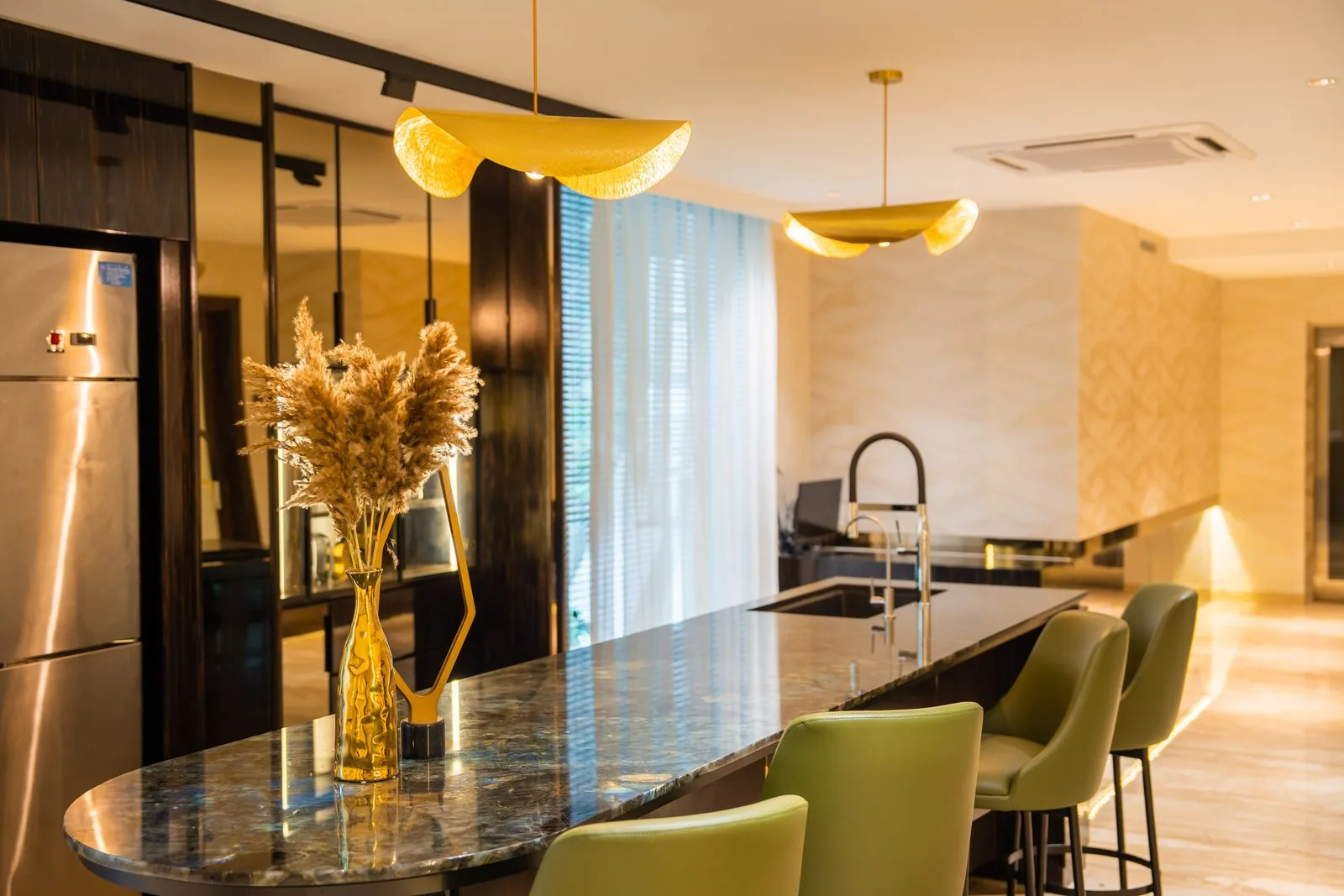Curved kitchen island with a polished stone countertop, olive bar stools and gold leaf pendants in a Bangsar bungalow