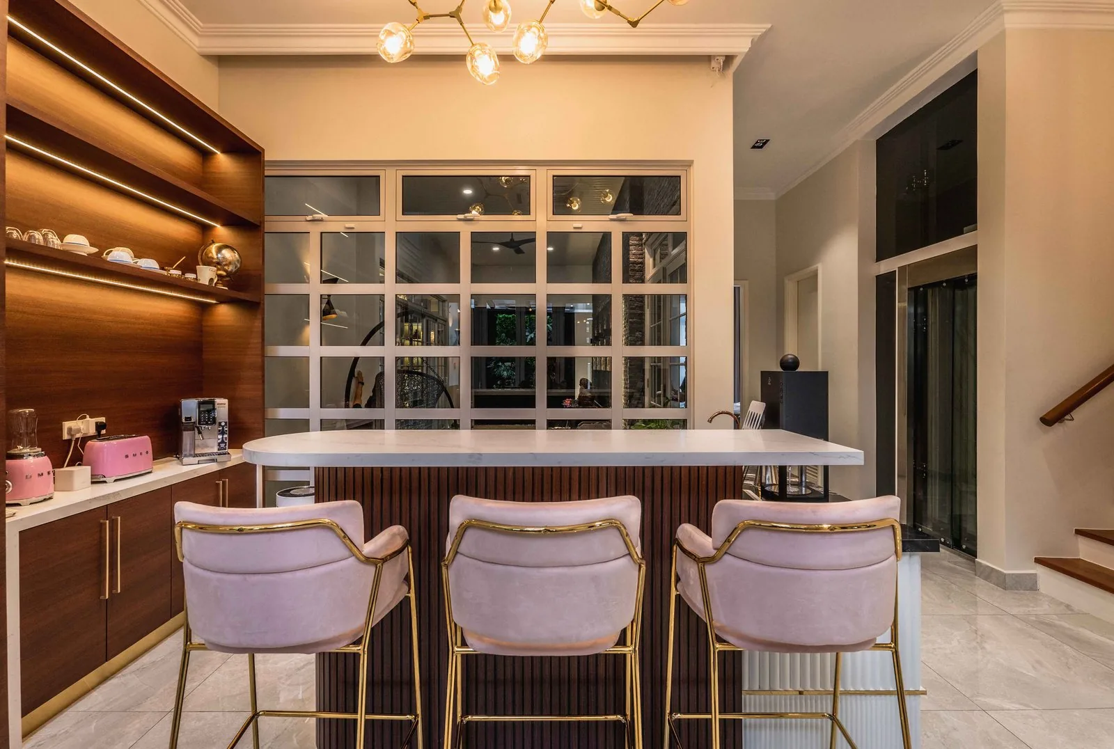 Kitchen island breakfast bar with pink stools in front of lit timber shelving at EcoSpring Villa