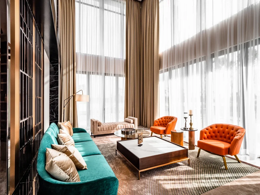 Double-height living room with a teal velvet sofa, orange armchairs and full-height windows in a Malaysian bungalow designed by Blaine Robert Design